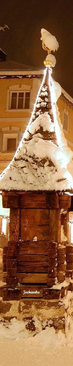 a small wooden structure covered in snow next to a building at night with lights on