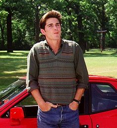 a man standing next to a red car in the grass with his hands on his hips