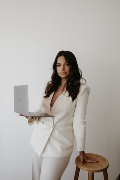 a woman in a white suit is holding a laptop and standing next to a stool