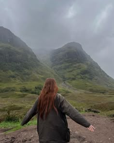 a woman standing on top of a dirt road in front of green mountains with her arms outstretched