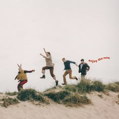 four people jumping in the air on top of a sandy hill with grass and sand