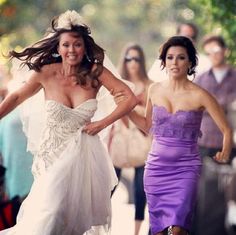 two women in dresses are walking down the street with other people behind them and one woman is wearing a purple dress