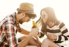 a man and woman sitting on the beach, one holding a beer bottle in his hand