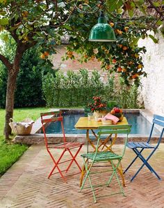 an outdoor table and chairs with fruit on it in front of a swimming pool surrounded by greenery