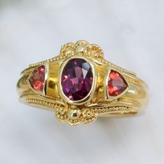 Wrapped in a luxurious setting of 18K granulated gold, our elegant Garnet & Sunstone ring offers a captivating display of gorgeous fall colors. The striking centerpiece is a beautiful rhodolite garnet stone, set off by two sunset-hued sunstones that sparkle with subtle warmth. This exquisite piece will add timeless sophistication to any wardrobe. 18KY granulated ring with 7x5mm rhodolite garnet and 2- 4mm trillion cut sunstones. The granulation on runs part way down the band resulting in a band Gold Ruby Birthstone Ring With Gemstone Accents, Yellow Gold Ruby Ring With Tourmaline Center Stone, Gold Garnet Birthstone Ring With Center Stone, Heirloom Ruby Ring With Gemstone Accents, Gold Heirloom Ruby Ring With Gemstone Accents, Heirloom Gold Ruby Ring With Gemstone Accents, Yellow Gold Garnet Rings With Gemstone Accents, Elegant 22k Gold Ruby Ring, Gold Garnet Multi-stone Ring