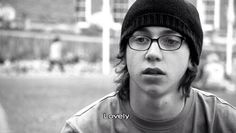 a young man wearing glasses and a beanie looks up at the camera while standing in front of a building