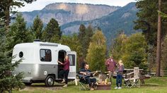 three people sitting in chairs next to an airstream with mountains in the back ground