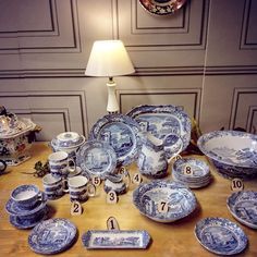 a table topped with lots of blue and white plates next to a lamp on top of a wooden table