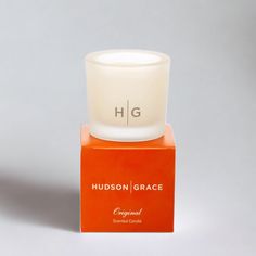 a white candle sitting on top of a red box next to a gray background with the words hudson grace written across it