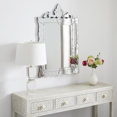 a white table with a lamp and mirror on it next to a vase filled with flowers