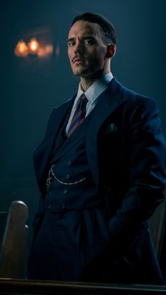 a man wearing a suit and tie standing in front of a chair with his hands on his hips