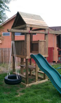 a wooden play structure with a slide and swing set in the grass next to a house