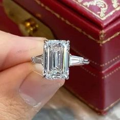 a woman's hand holding an emerald and baguette ring in front of a red box