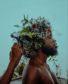 a man with flowers on his head standing next to another man's face and looking up at the sky