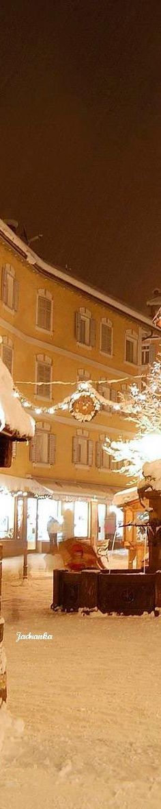 a snow covered park bench sitting in front of a tall building at night with lights on it