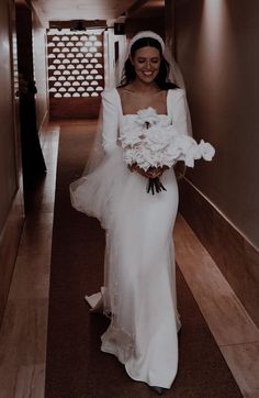 a woman in a wedding dress walking down a hall way holding a bouquet of flowers