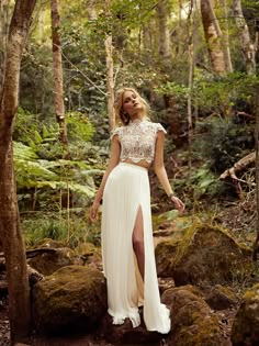 a woman standing in the woods wearing a white skirt and crop top with high slit