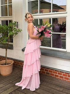 a woman in a long pink dress holding flowers by a window with potted plant