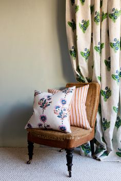 a chair with two pillows on it in front of a window covered by drapes