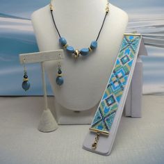 The good look of turquoise and gold is what makes this set so becoming. This bracelet/necklace/earrings set has a geometric design of diamond shapes. They are handmade with care using a bead loom to make the bracelet. Delica beads were used giving the bracelet a more polished and uniform look. The bead colors are silver lined teal, matte cyan turquoise blue, chrysoprase (light green), butter rum and white. The bracelet's pattern has a diamond shaped center with four diamond shapes inside and chevrons on the outside with a teal X shape. The ends have basically the same, diamonds with a teal X shape adding an arrow shape. It has gold plated tube ends and an extension chain of 1 1/2 inches for sizing... or to loosen the fit, simply hook into the chain. From the lobster claw clasp to the ring Elegant Adjustable Turquoise Jewelry Set, Adjustable Bohemian Blue Jewelry Sets, Handmade Geometric Gold Jewelry, Gold Beaded Geometric Jewelry, Geometric Beaded Gold Jewelry, Gold Geometric Beaded Jewelry, Adjustable Beaded Blue Jewelry Set, Geometric Gold Beaded Jewelry, Blue Bohemian Jewelry With Geometric Shape