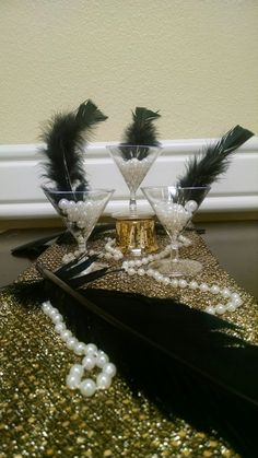 two martini glasses with black feathers and pearls on a gold sequin covered table cloth