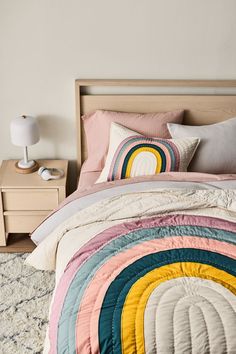 a bed with a rainbow quilt on top of it next to a night stand and lamp