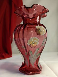 a red glass vase with flowers painted on the bottom and sides, sitting on a white surface