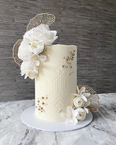 a white wedding cake with flowers and gold accents on a marble countertop in front of a gray wall