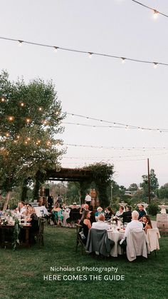 a group of people sitting around a table on top of a lush green field with string lights