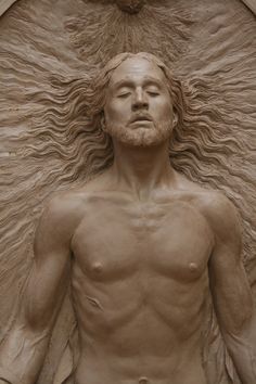 a man with long hair and beard standing in front of a wall sculpture that looks like he has his eyes closed