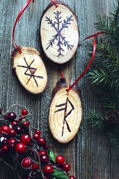 three wooden ornaments with designs on them sitting next to berries and pine cones, one has a red ribbon around it