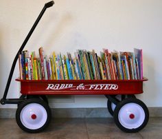 a red radio flyer wagon with books on the top and white wheels, in front of a wall