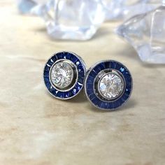 This pair of vintage style stud earrings features round brilliant cut diamonds with a sapphire halo. The total approximate diamond weight is 1.00 carats with J color and VS clarity. These earrings are platinum, with push-backs. ✦ DIAMOND SPECIFICATIONS: Diamond Cut: Brilliant Cut Diamond Weight 1.00 Carats Total ✦ ENGAGEMENT RING SPECIFICATIONS: Ring Material: Platinum Stones: Diamond ✦ WHAT COMES IN YOUR SHIPMENT: - Your Engagement Ring - Quality Ring Box - Jewelry Cleaner - UGL Certificate ✦ W Luxury Sapphire Diamond Earrings With Halo Design, Classic Sapphire Earrings For Anniversary, Classic Sapphire-colored Cubic Zirconia Diamond Earrings, Classic Sapphire Diamond Earrings With Halo Setting, Classic Sapphire Earrings With Halo Setting, Sapphire Diamond Earrings Fine Jewelry, Sapphire Diamond Earrings For Wedding, Gia Certified Blue Diamond Round Earrings, Vintage Round Platinum Diamond Earrings