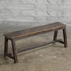 an old wooden bench sitting in front of a white brick wall on concrete flooring