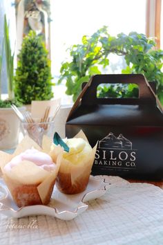 two cupcakes sitting on a plate in front of a black jug and potted plants