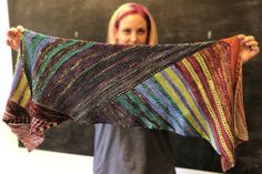 a woman holding up a multicolored shawl in front of a chalkboard