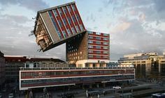 an upside down building in the middle of a parking lot with cars and people walking around