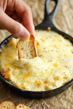 a hand dipping a piece of bread into a skillet filled with cheesy cheese