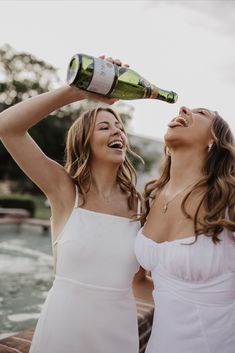 two beautiful women standing next to each other and drinking from bottles in front of the water