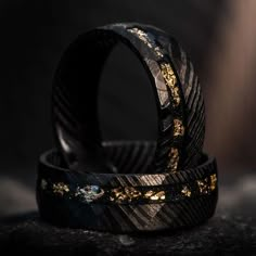 two black and gold wedding bands sitting on top of each other in front of a dark background