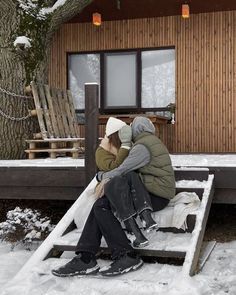 two people sitting on a bench in front of a house covered in snow and sleds