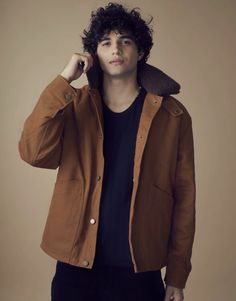 a man with curly hair wearing a brown jacket and black t - shirt is posing for the camera