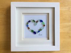 a heart made out of sea glass sitting in a white frame on a wooden table