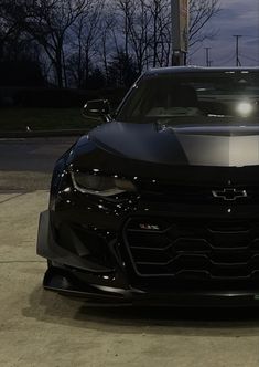 the front end of a black sports car parked in a parking lot at night with its lights on