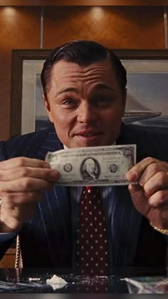 a man in a suit and tie is holding up a $ 1 bill while sitting at a desk