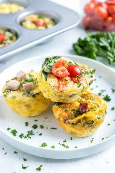 three small muffins on a white plate with tomatoes and other food items in the background