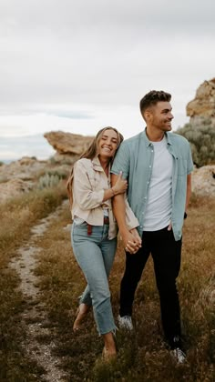 a man and woman holding hands while walking through the grass in front of some rocks