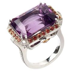 A splendid ring 18kt white gold showcases a mesmerizing center emerald-cut amethyst, which exudes an air of regal elegance. Encircling this captivating amethyst are vibrant orange round sapphires, creating a striking contrast that adds a touch of warmth and drama. To complete this exquisite composition, diamonds adorn the sides of the ring, adding a brilliant sparkle that complements the gemstones. Ring Emerald Cut Amethyst 12.52 cts. with a Halo of 24 Orange Sapphires 1.28 cts., Diamonds 0.92 cts., and 2 trillion Sapphires 0.11 cts. in 18kt White Gold. Size 5.75 Perfect gift for Christmas, Mom, Girlfriend, Daughter, Graduation, Birthday and more. Daughter Graduation, Ring Emerald Cut, Round Sapphire, Orange Sapphire, Ring Emerald, Christmas Mom, Vibrant Orange, Gift For Christmas, Cocktail Rings