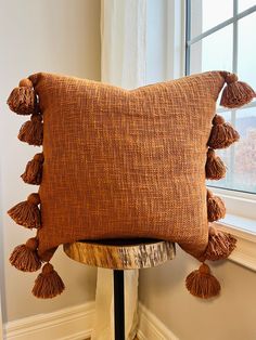 a brown pillow with tassels sitting on top of a wooden table next to a window