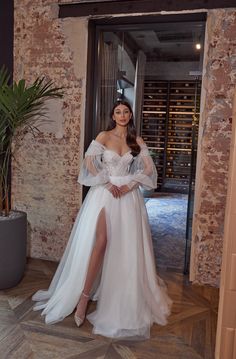 a woman standing in front of a doorway wearing a white dress with sheer sleeves and thigh high slit